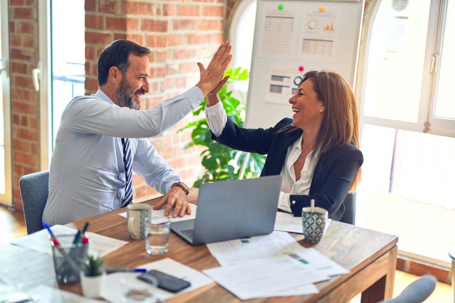 Two business professionals smiling and high-fiving at a desk after completing a project, representing local business services in Tampa including consulting, digital marketing, print marketing, and coaching